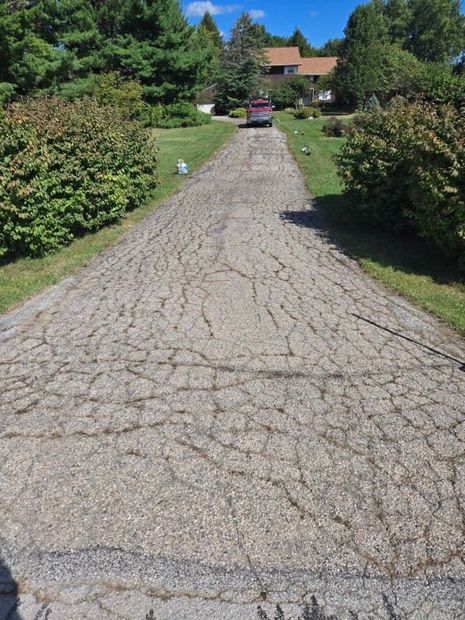 A cracked asphalt driveway leads to a house; grass and bushes line the sides. Blue sky and trees in the background.