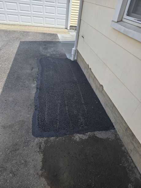 Asphalt driveway with a rectangular patch of fresh asphalt next to a light yellow building.