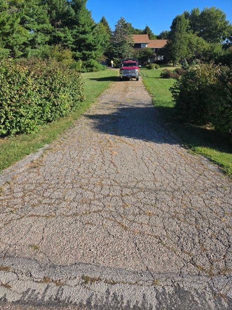 Gravel driveway leads to a house; red truck parked at the end. Green bushes on each side.