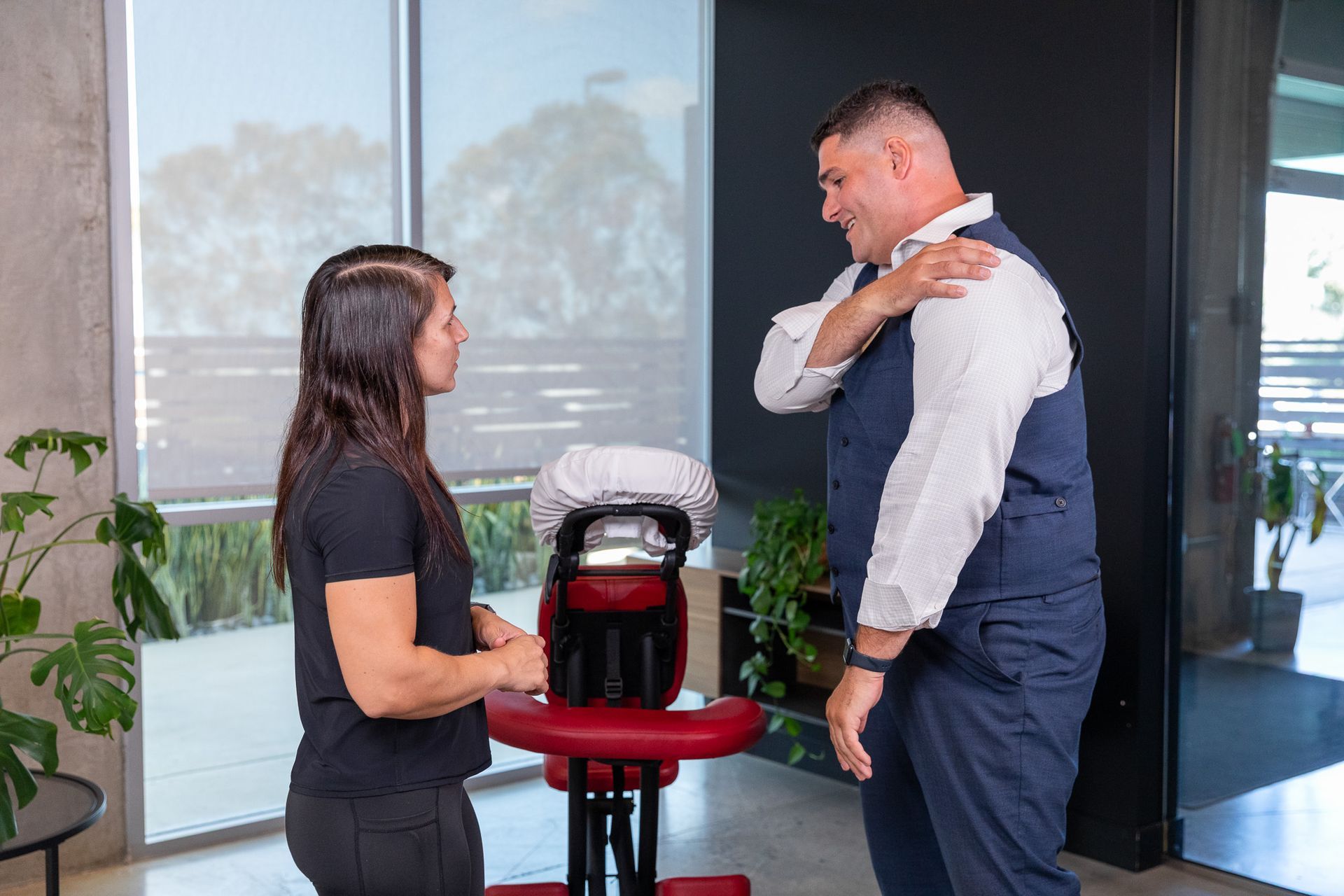 Woman in black shirt consults man in vest, who points to his shoulder near a massage chair.