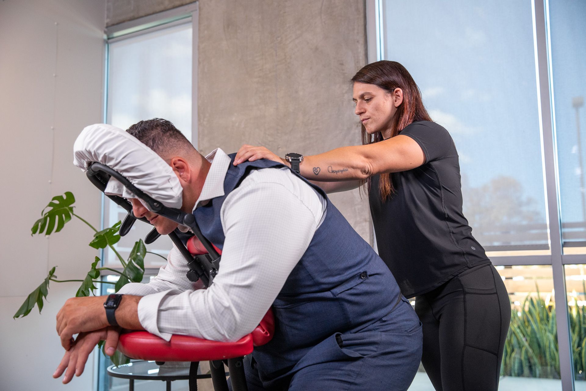 Woman giving a massage to a person in a chair; indoor setting with plants.