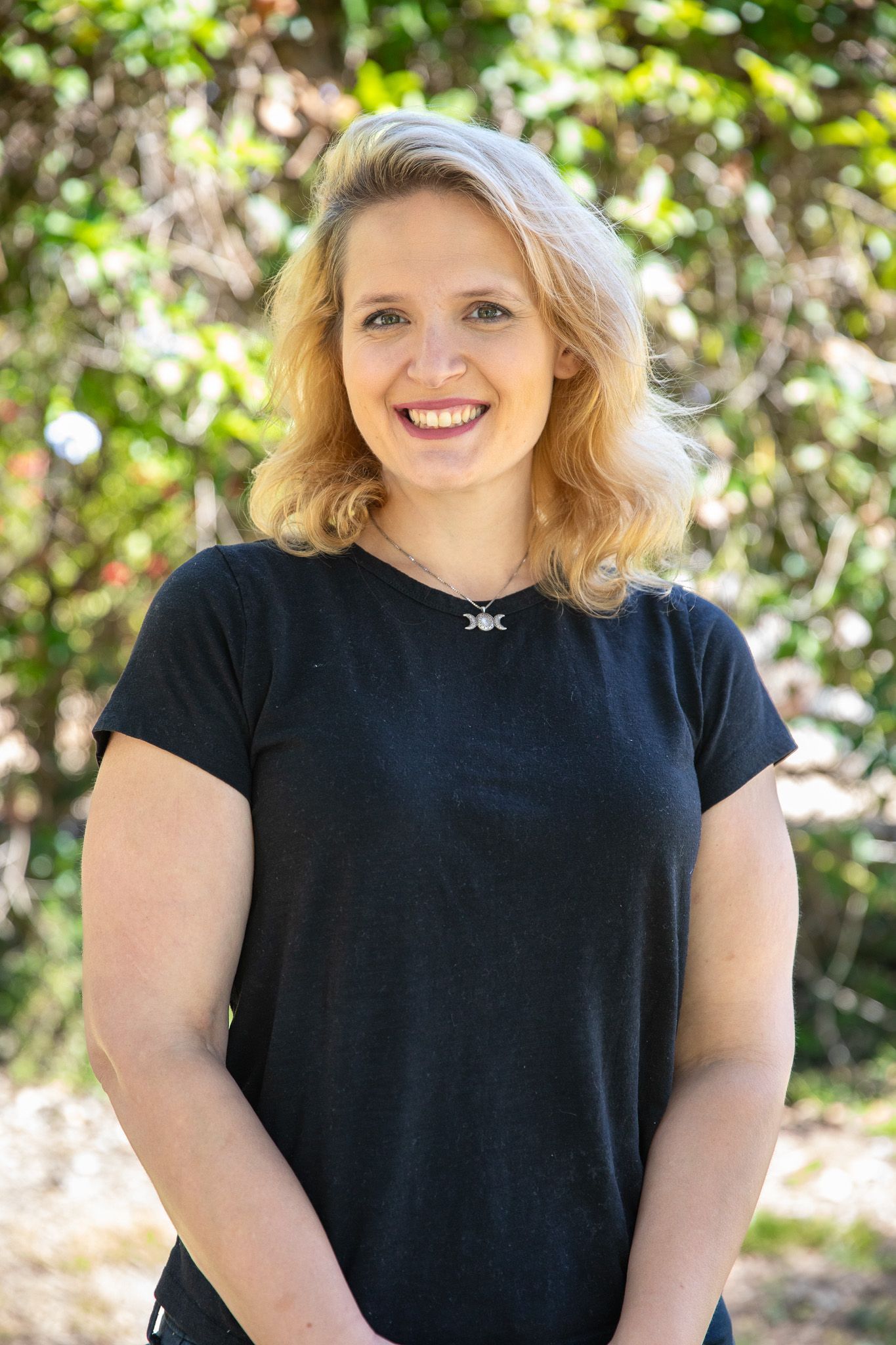 Woman with blonde curly hair, wearing a black shirt, smiling outdoors.