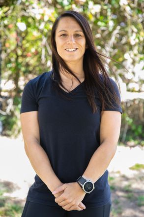 Woman with dark hair, wearing a black v-neck shirt and watch, smiles outdoors.