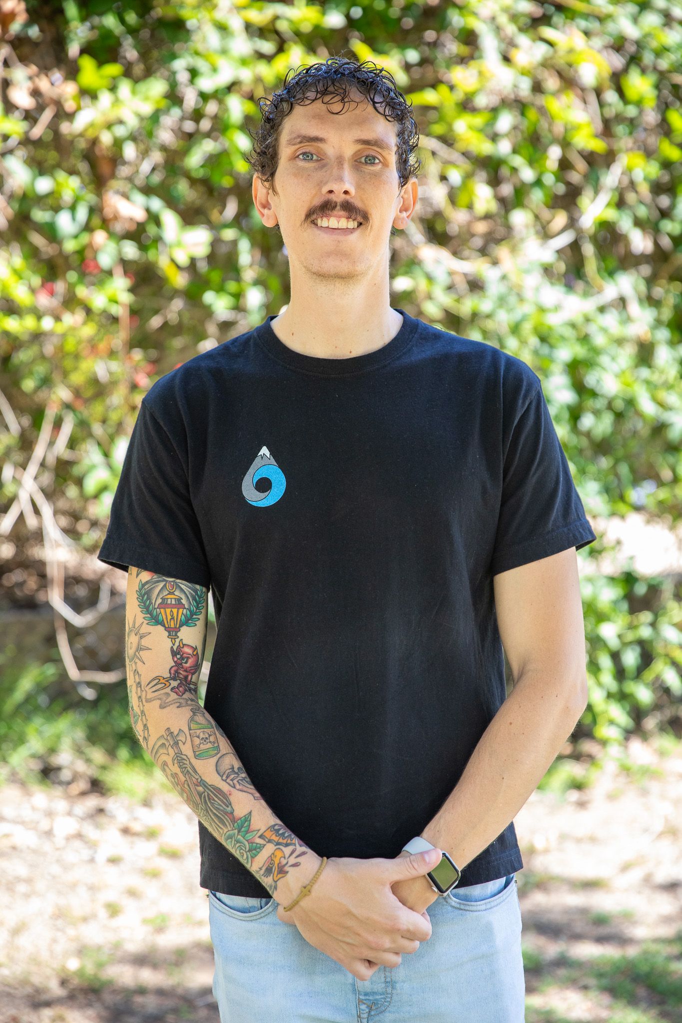 Man with tattoos, mustache, and black shirt in front of trees, hands clasped, smiling.