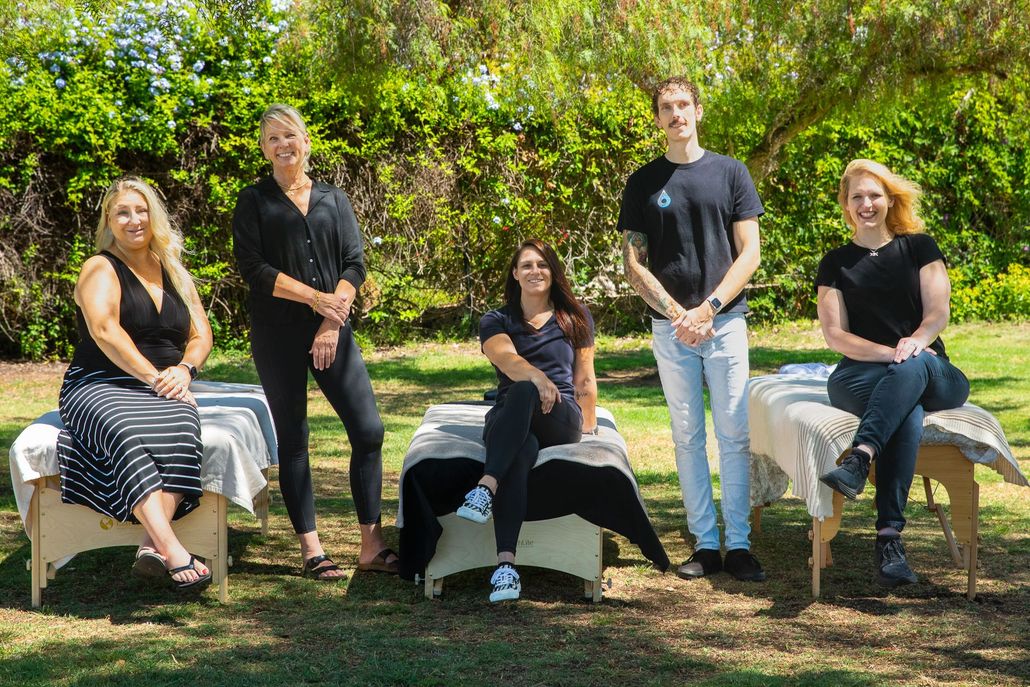 Five massage therapists pose outdoors with massage tables.