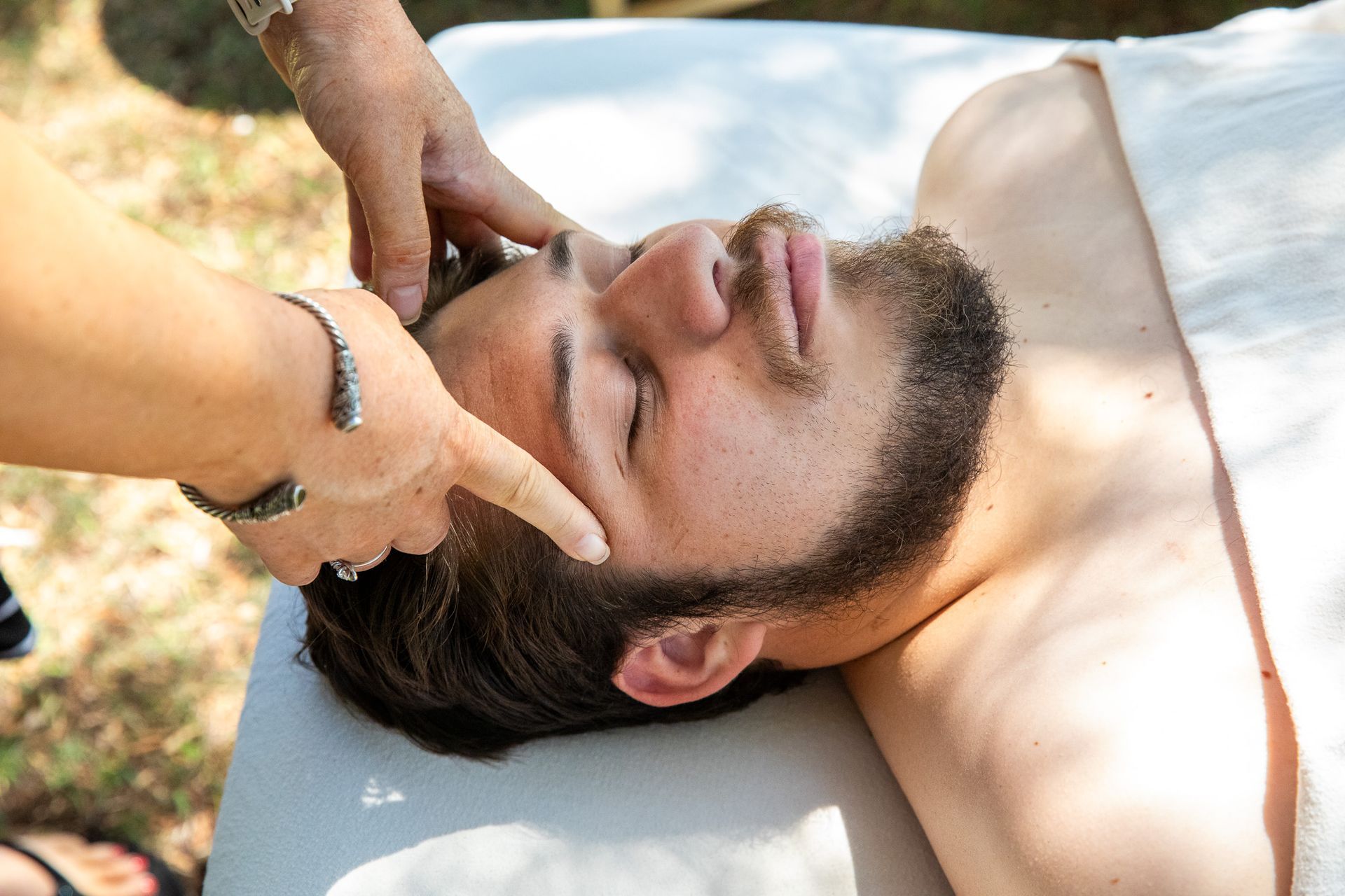 Person receiving a head massage outdoors; masseuse's hands on forehead, eyes closed.