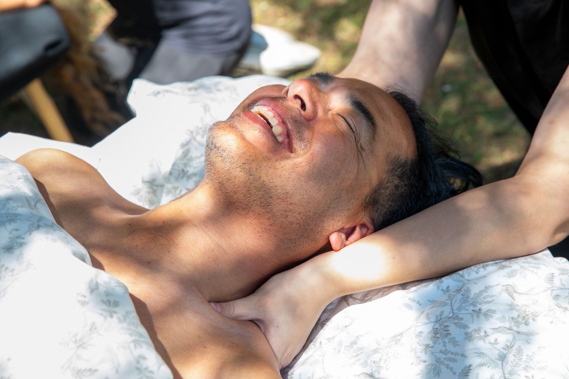 Man receiving a neck massage outdoors, smiling with eyes closed; sunlight.