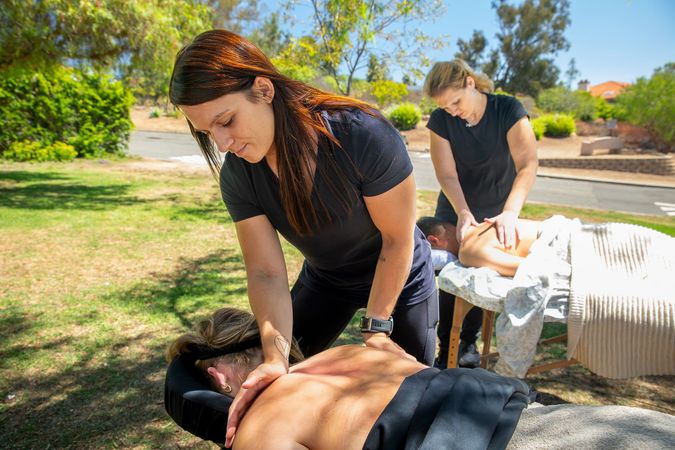 Two women giving back massages outdoors on a sunny day.