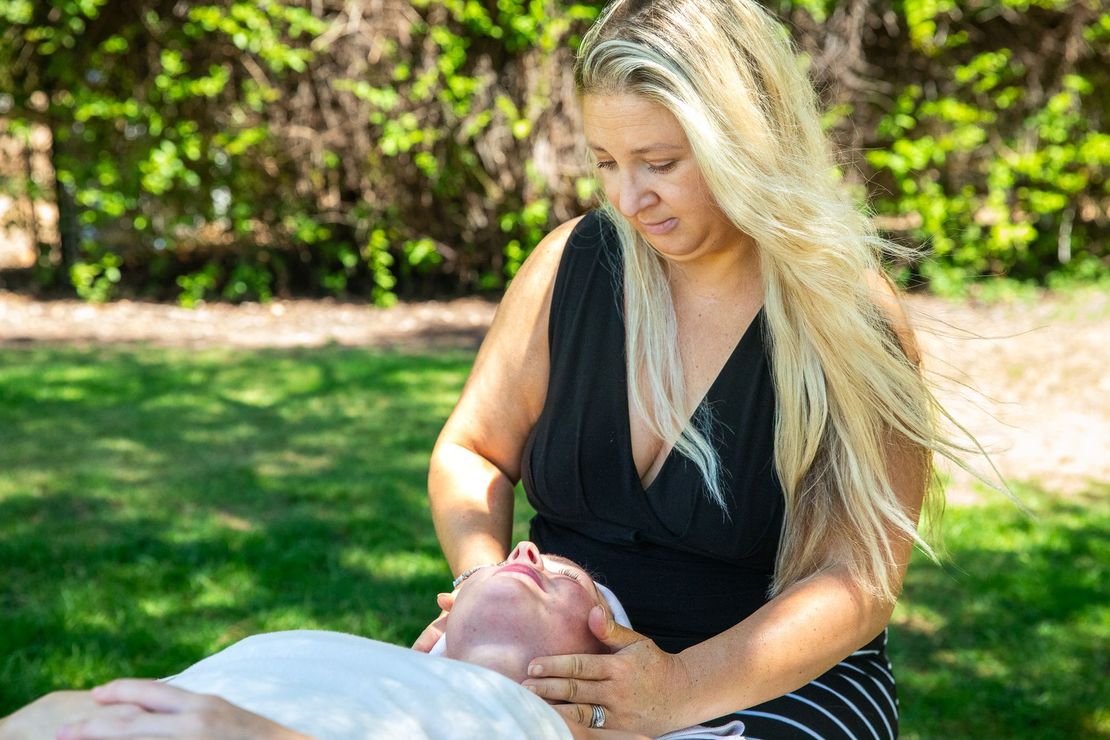 Woman giving facial massage outdoors; green grass, trees in background.