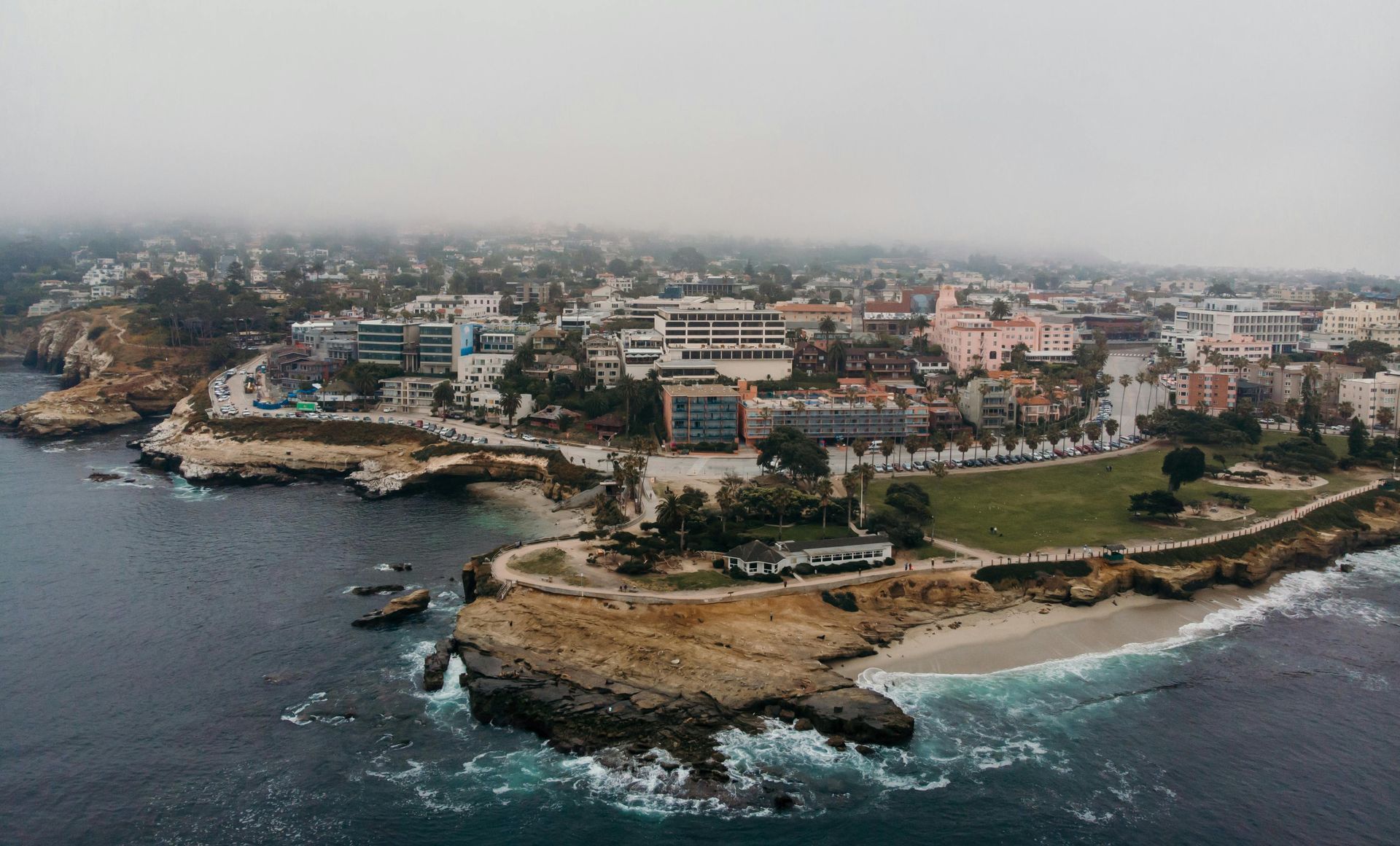 Coastal city with buildings, park, and rocky coastline under a foggy sky.
