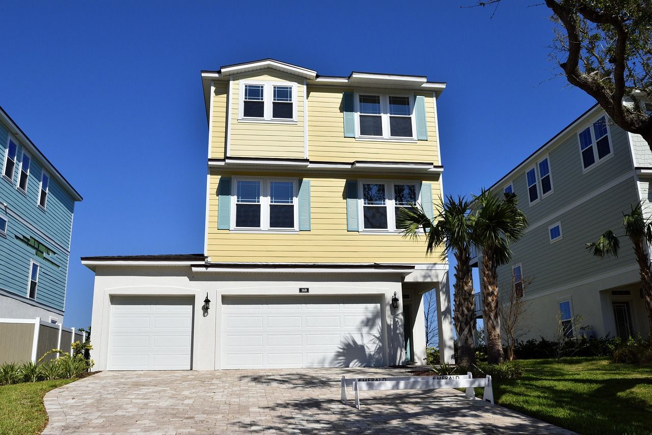A three-story yellow house with a two-car garage, flanked by neighboring houses under a clear blue sky.