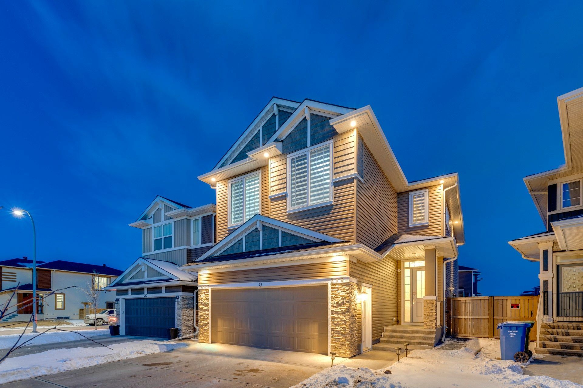 A multi-story suburban house with beige siding and stone accents, lit by exterior lights at dusk with snow on the ground.