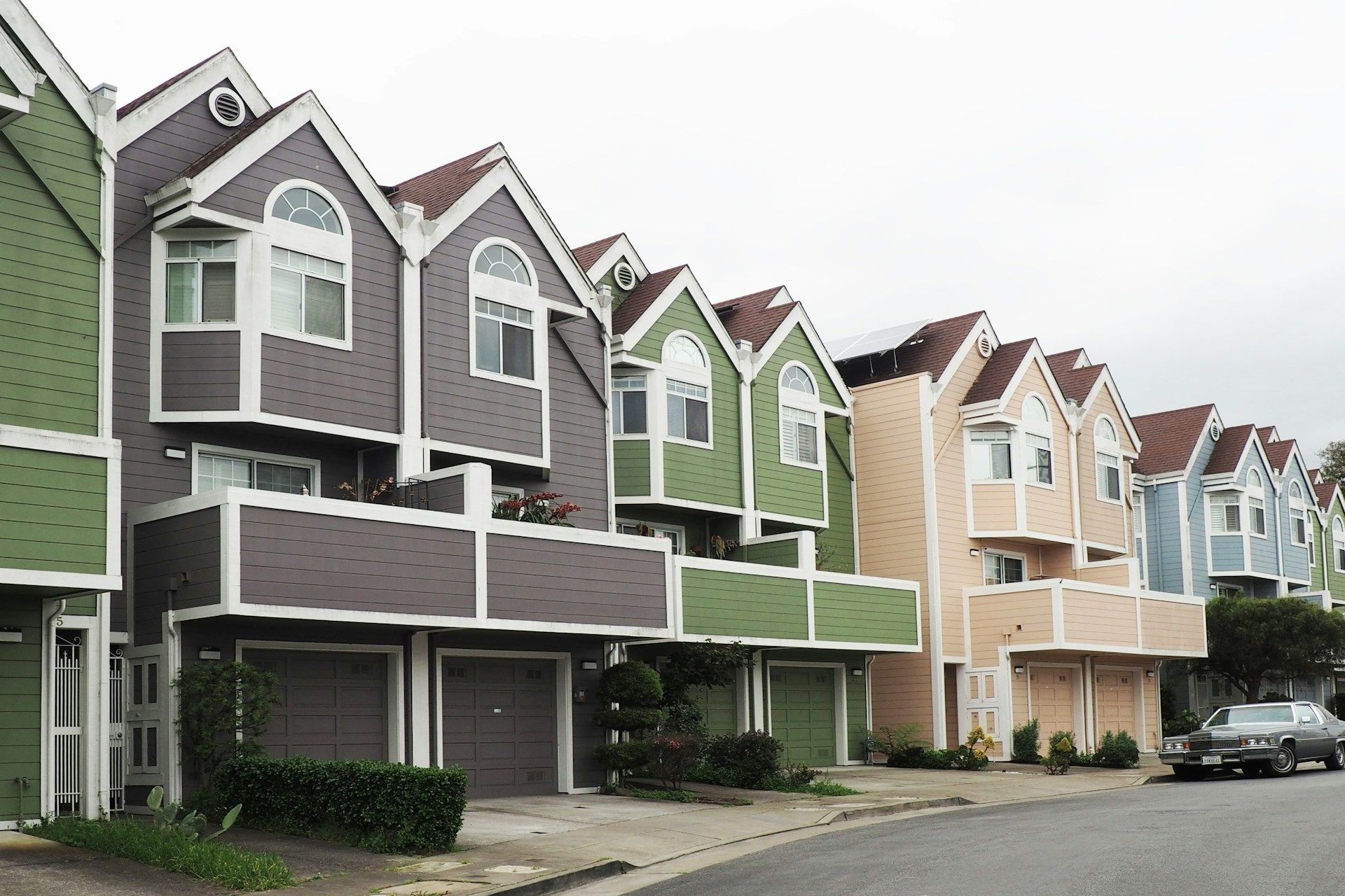 A row of multi-story townhouses with diverse paint colors, gabled roofs, and ground-level garages on a residential street.