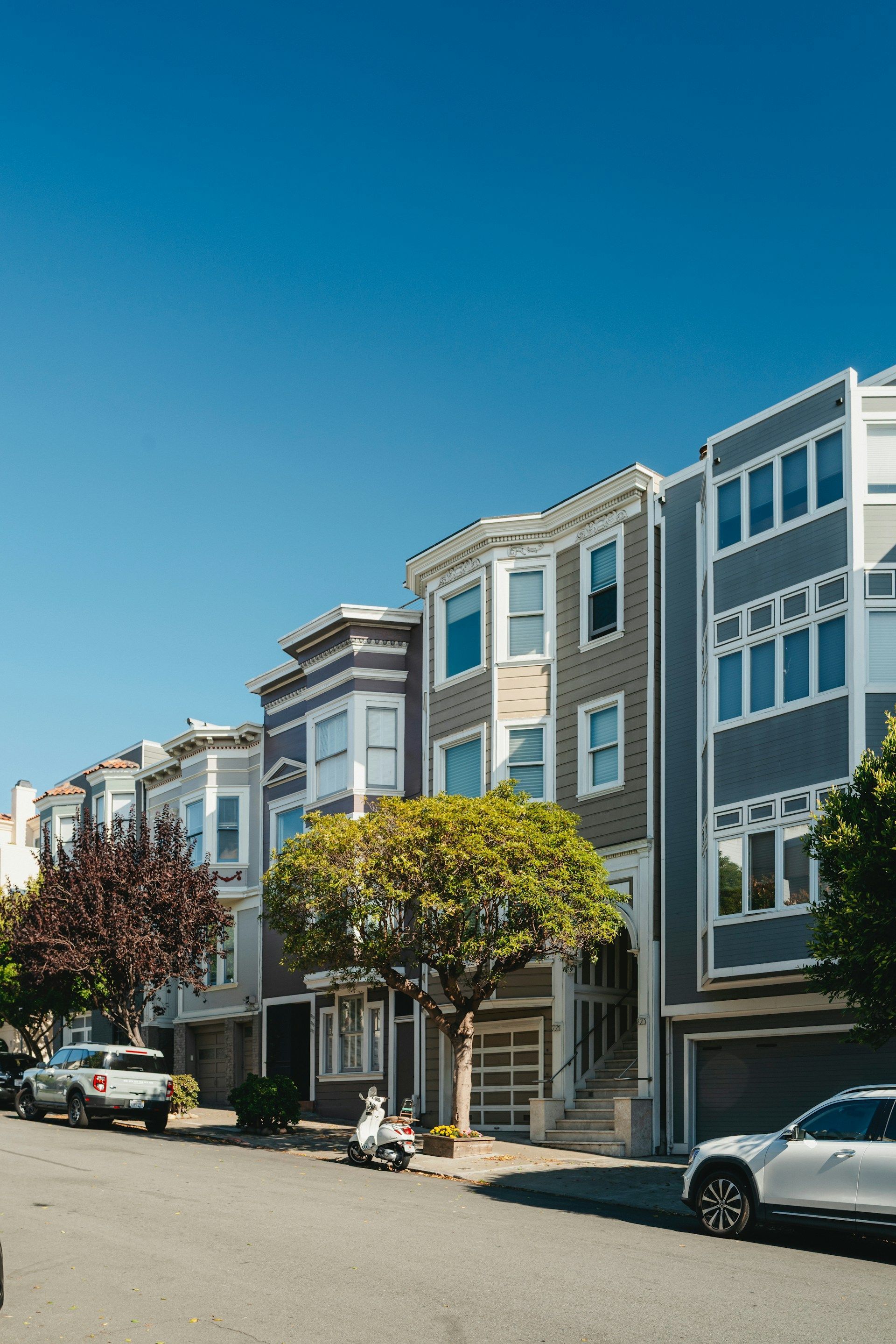 A street view of grey residential row houses on a sunny day with parked cars and a tree in the foreground.