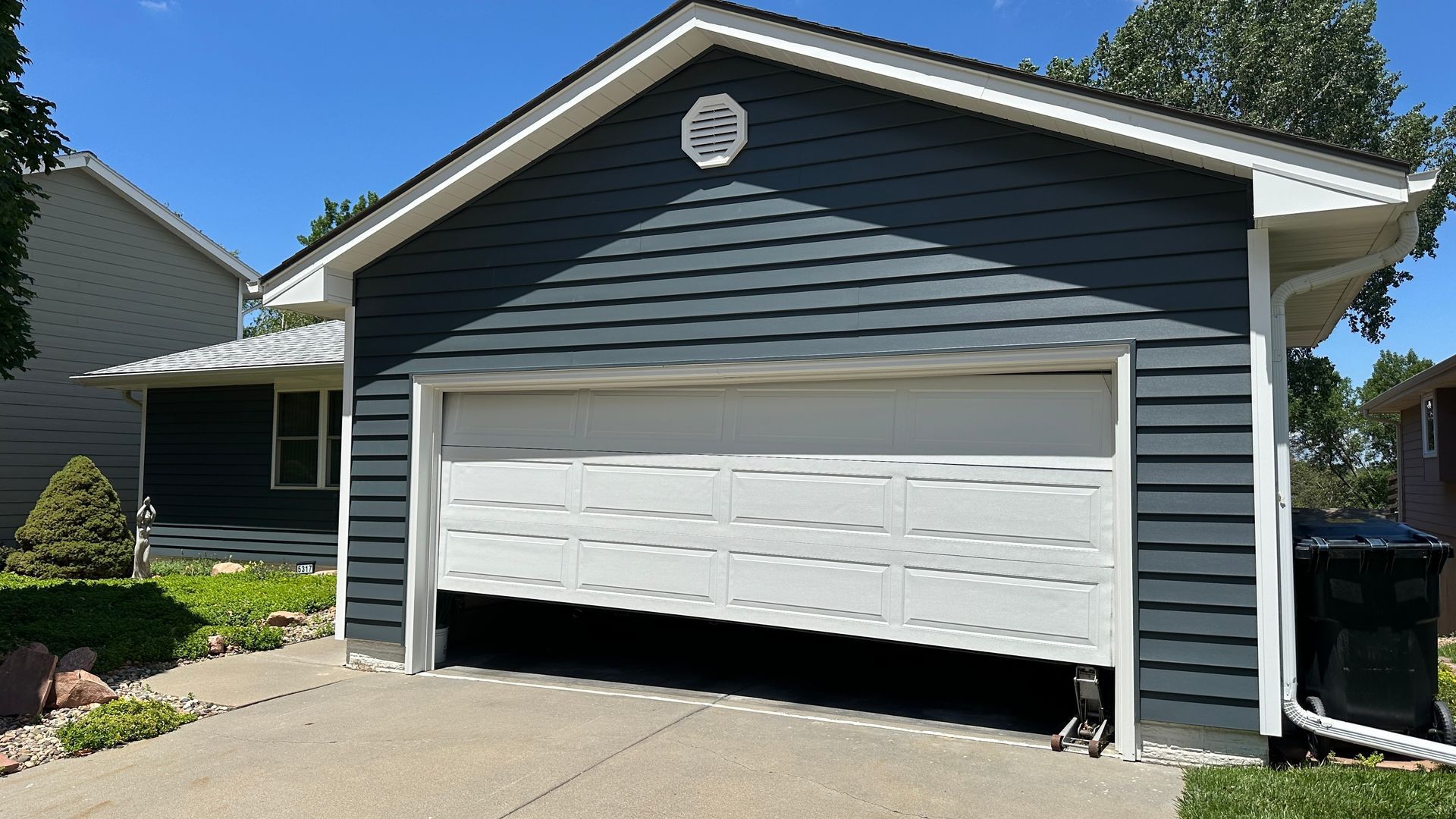 A hand holds a remote control to open a white garage door in front of a dark car on a red-walled building.