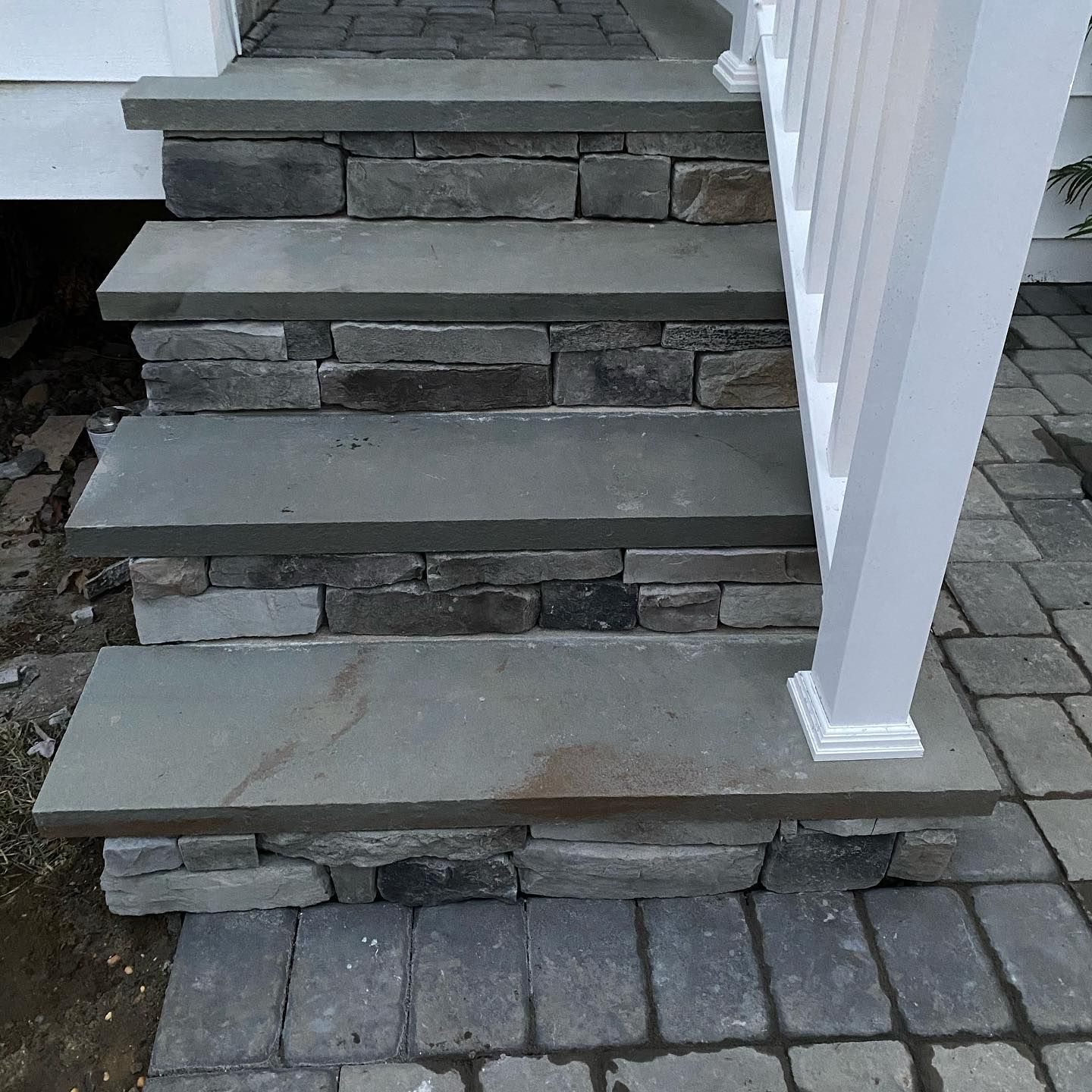 Stone steps leading up to a house entrance. White railing on the right. Brick pavers surround.