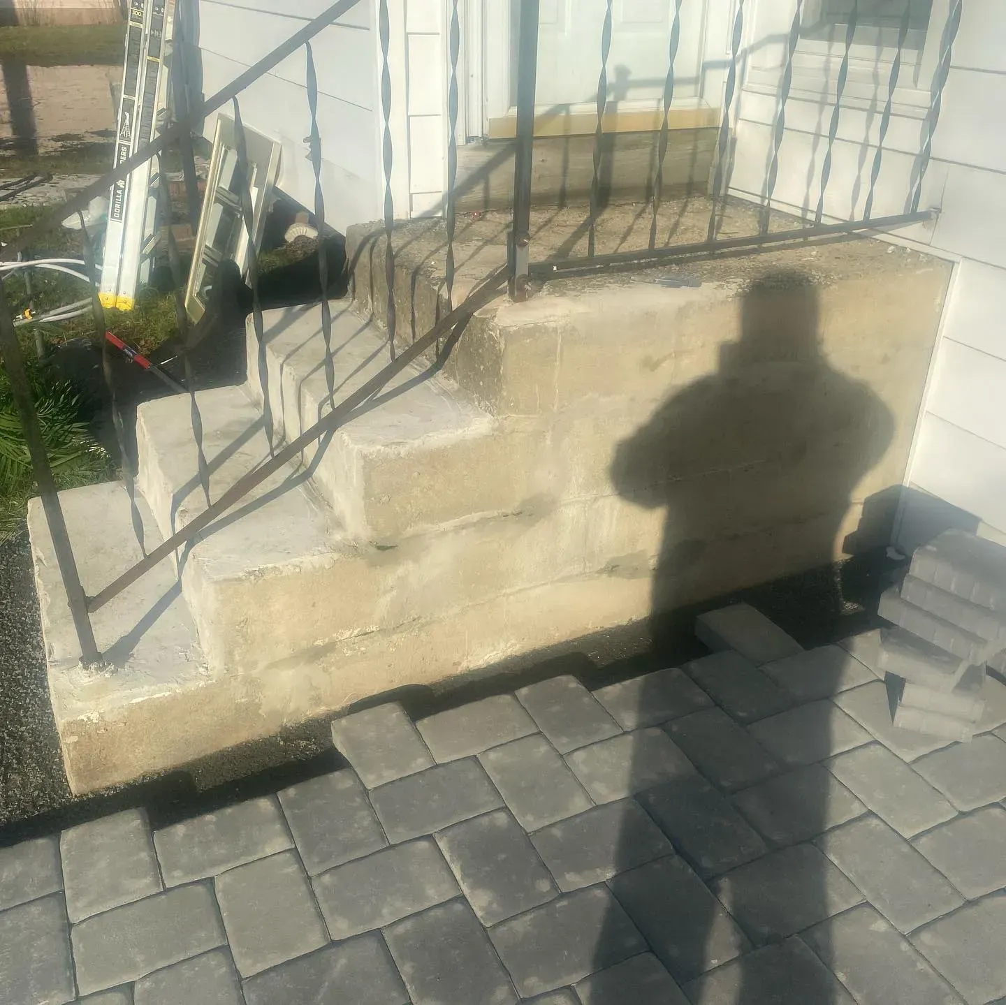 Concrete steps with black iron railing, being paved with gray brick. Shadow of person on steps.