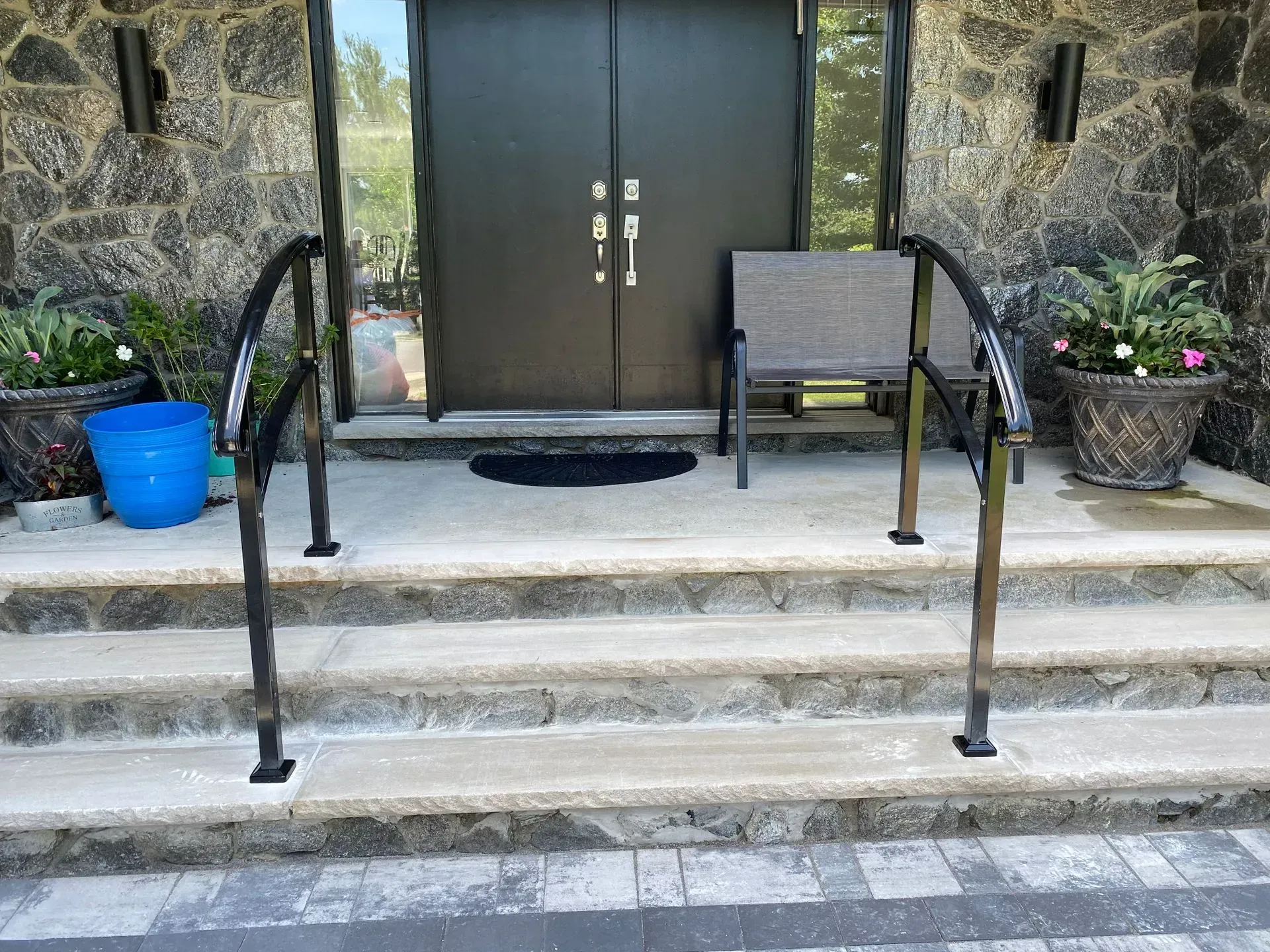 Exterior entrance with steps and black handrails, potted plants, and a chair.
