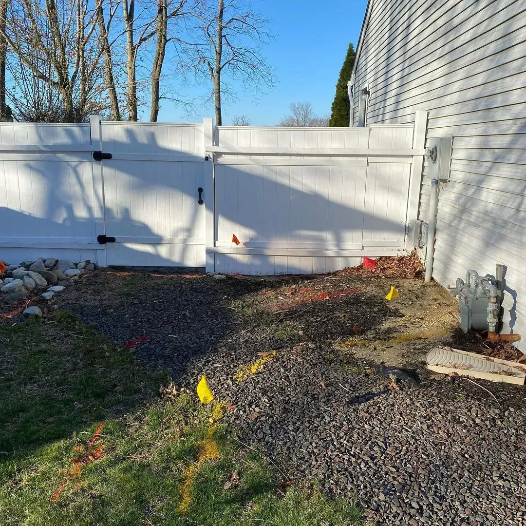 White fence with gate, rocky ground, yellow flags, and part of a house under blue sky.