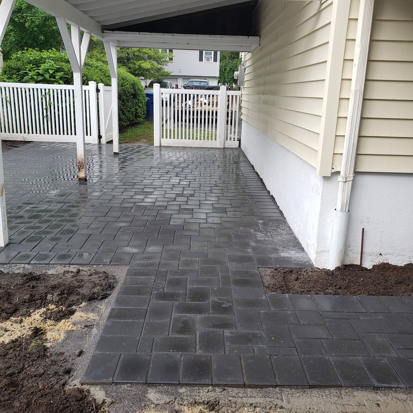 Dark pavers on a patio next to a house with a white fence and a covered area.