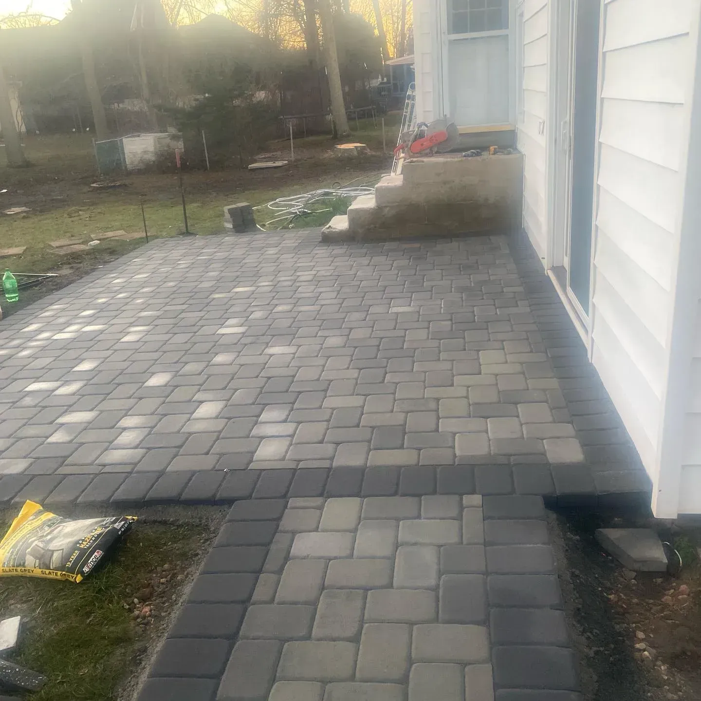 Patio paved with gray and black bricks, next to a white house with a set of stairs.