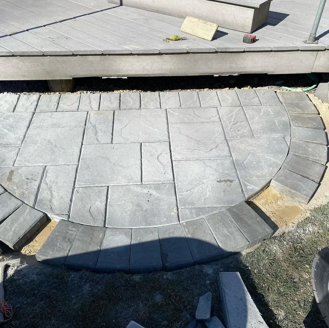 Asphalt paver patio in progress, semi-circle shape, under a deck. Gray and tan materials.