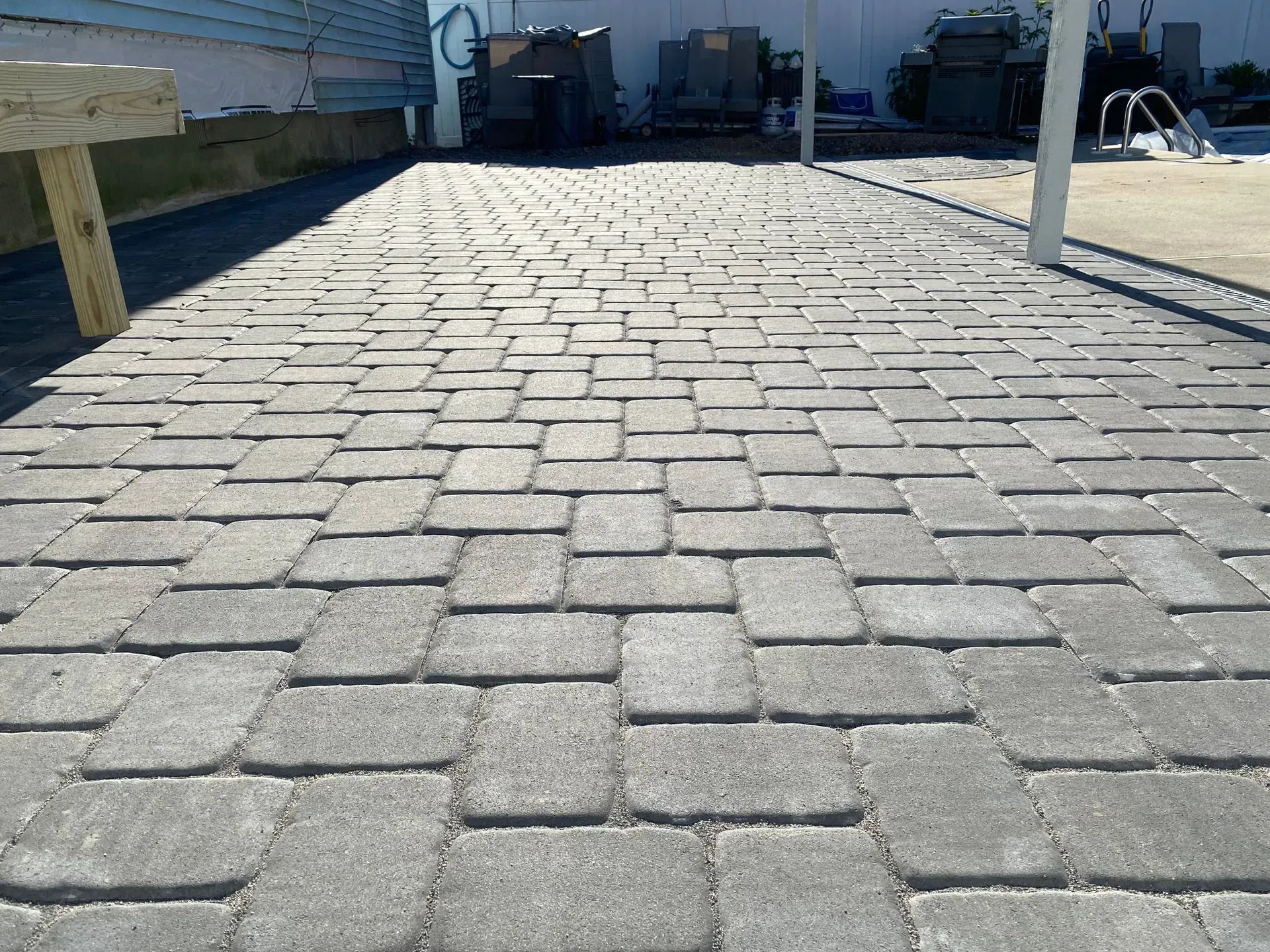 Gray brick pavers create a walkway, leading toward a background with equipment near a pool.