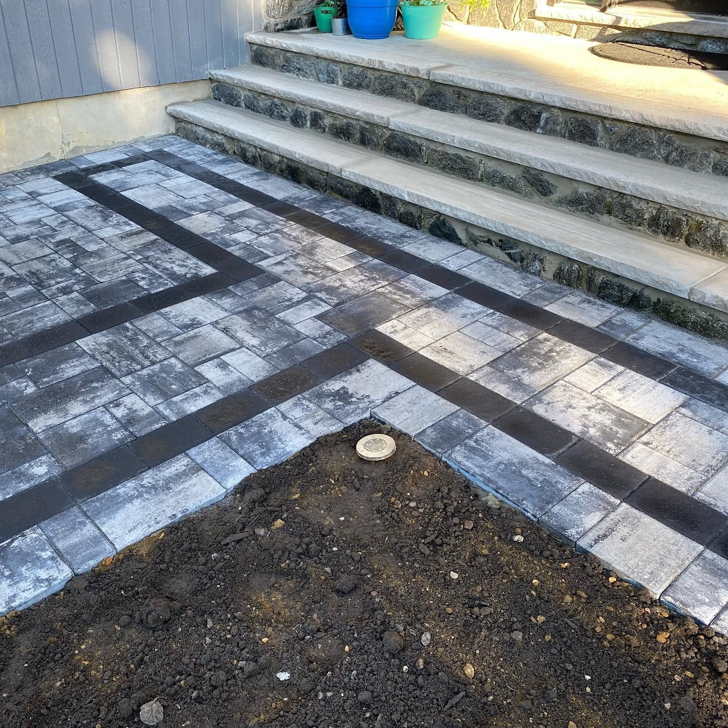 Paver patio with dark border next to stone steps. Unplanted soil in foreground.