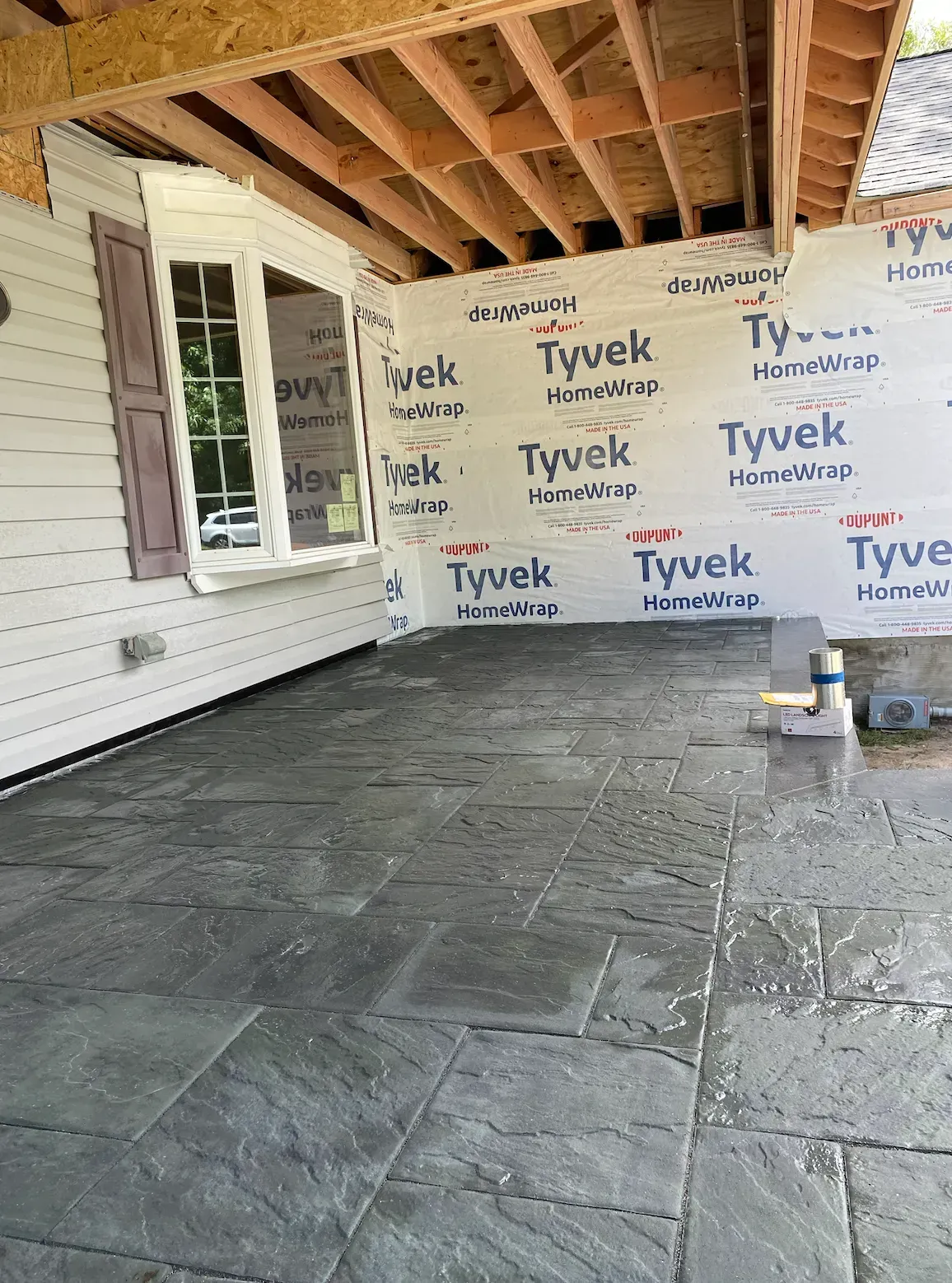 Patio under construction with gray stone tiles and a bay window, covered in Tyvek.