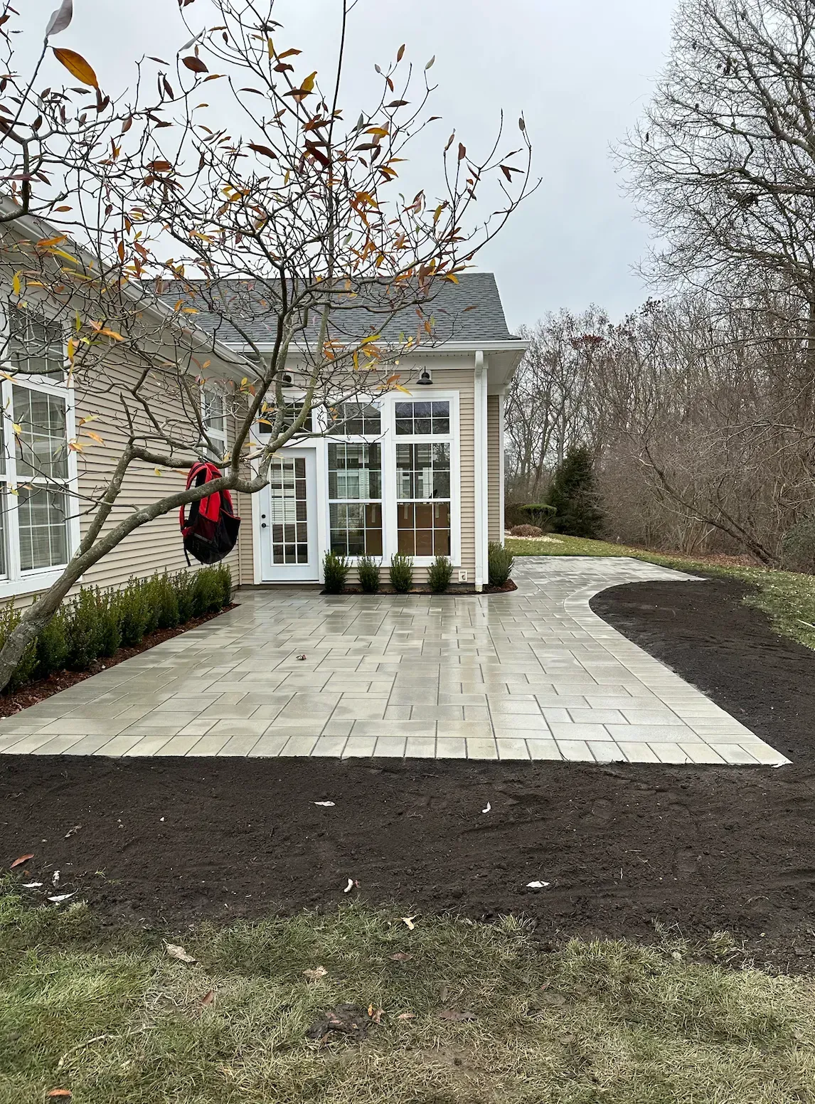 Brick patio extends from a house, bordered by mulch. A tree and shrubbery are in view.