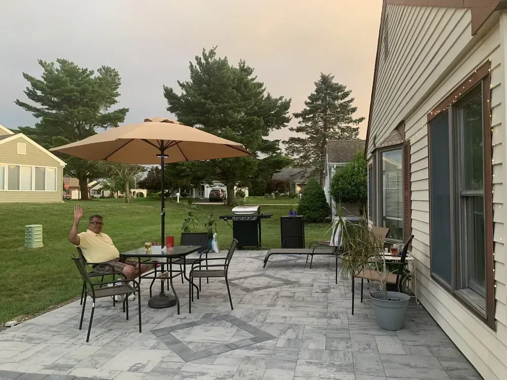 Man waving from a patio with table, umbrella, grill, and house, dusk setting.