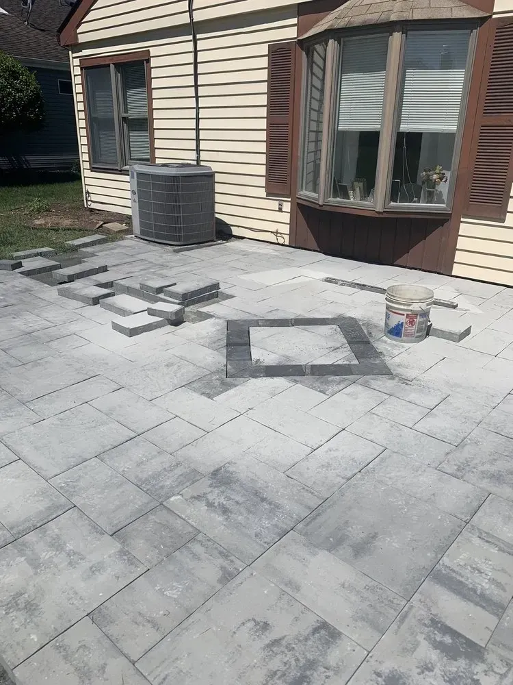 Patio under construction with gray pavers near a house with an air conditioner unit and bay window.