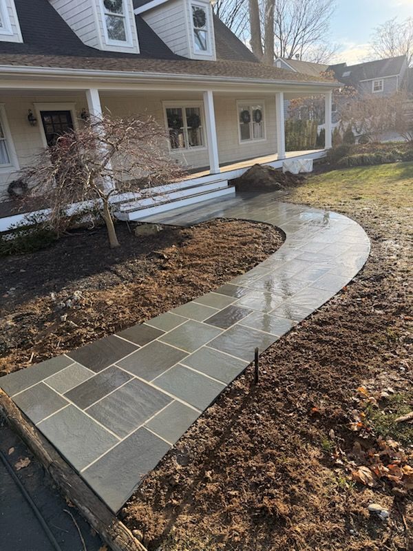 Stone walkway leading to a house with a porch. Dark gray rectangular pavers on a winding path, surrounding dirt and grass.