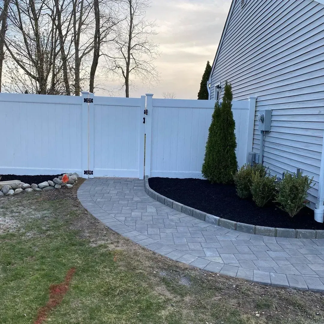 White fence with gate, brick pathway, and landscaping beside a house.