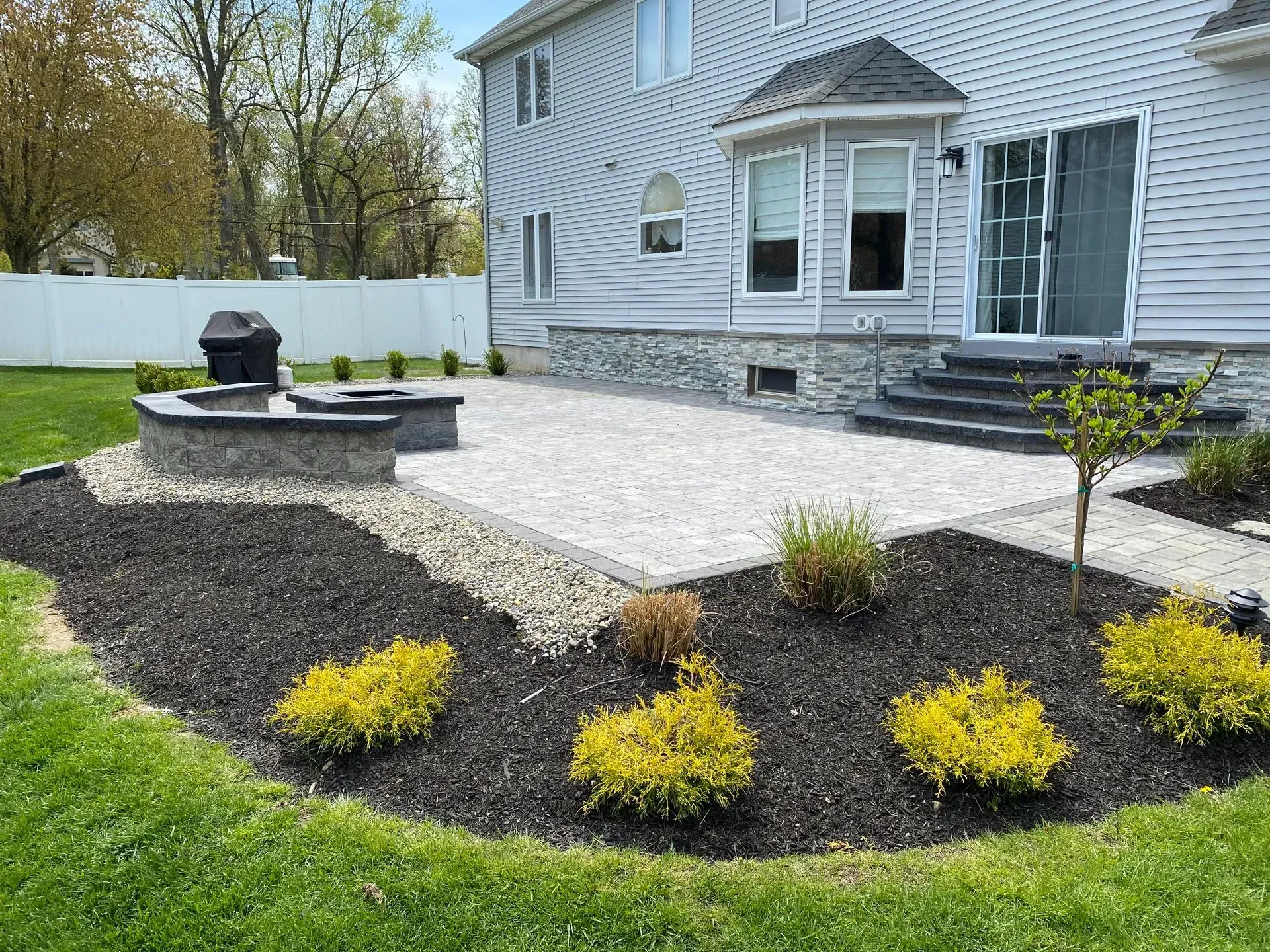 Backyard patio with fire pit, grill, and landscaping; house in background.