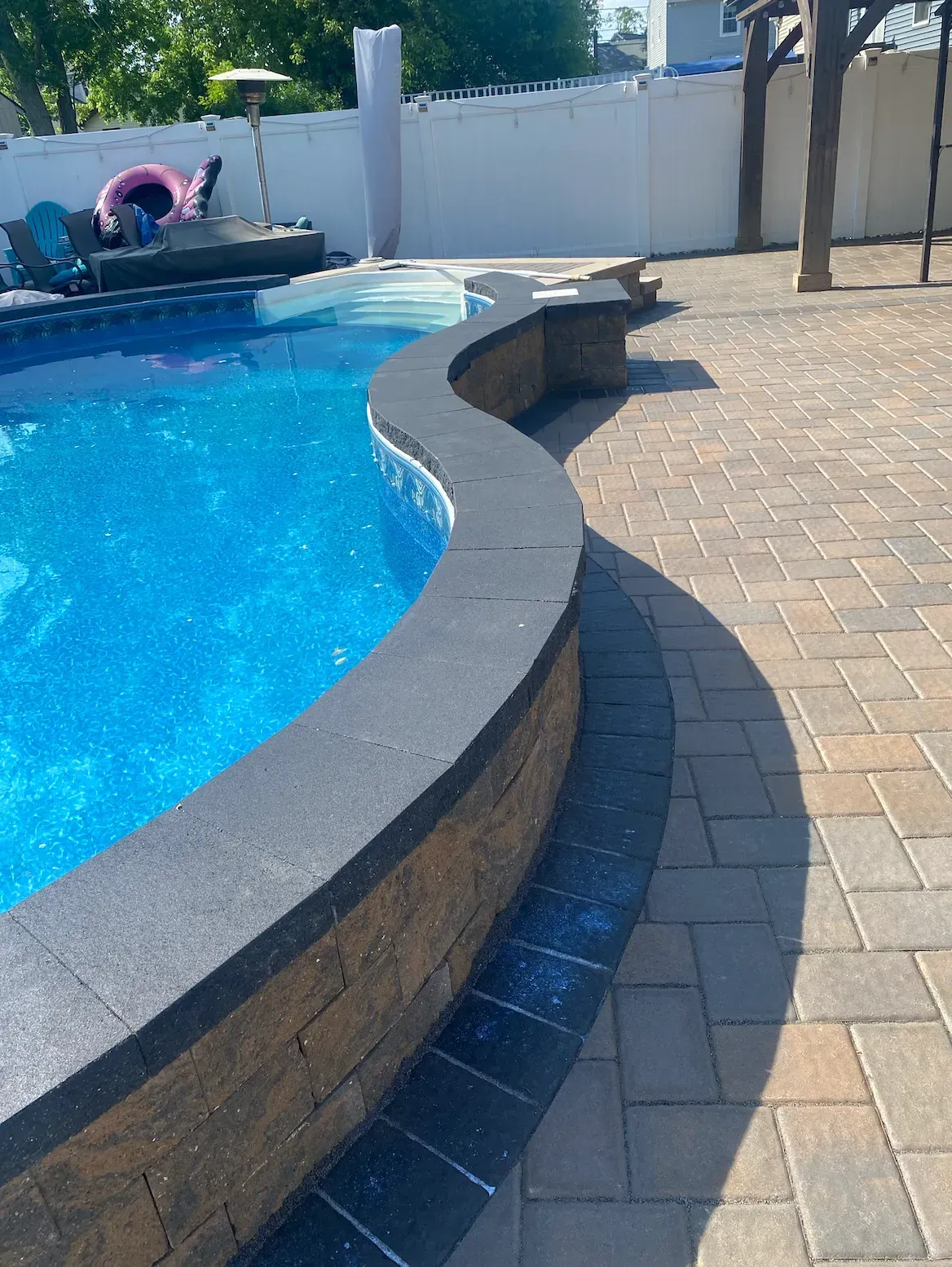A curved swimming pool with blue water, surrounded by brick patio. Sunny outdoor setting.