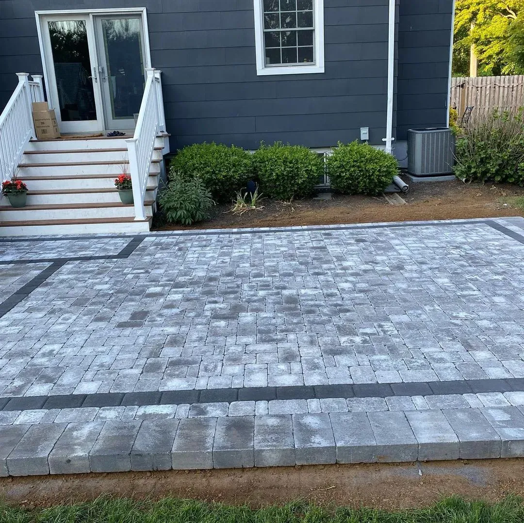Paver patio with dark gray house and white stairs in background.