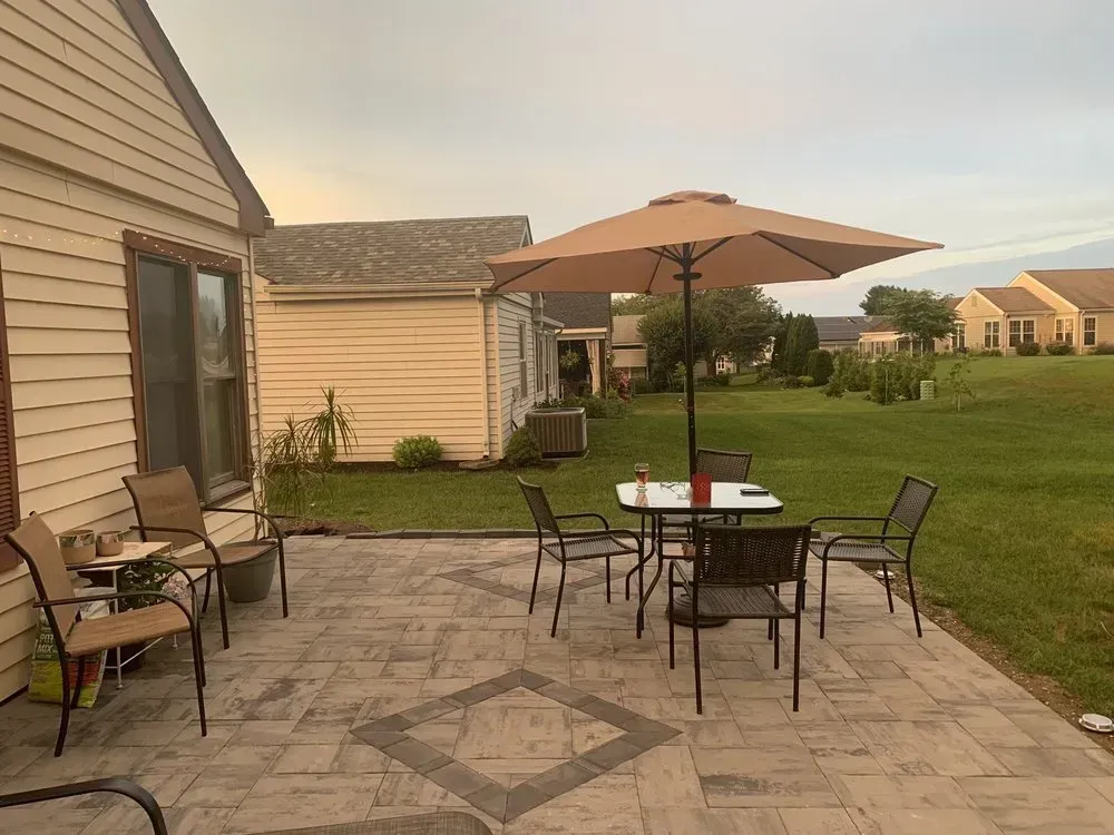 Patio with table, chairs, and umbrella; adjacent to a house. Green lawn and overcast sky in the background.