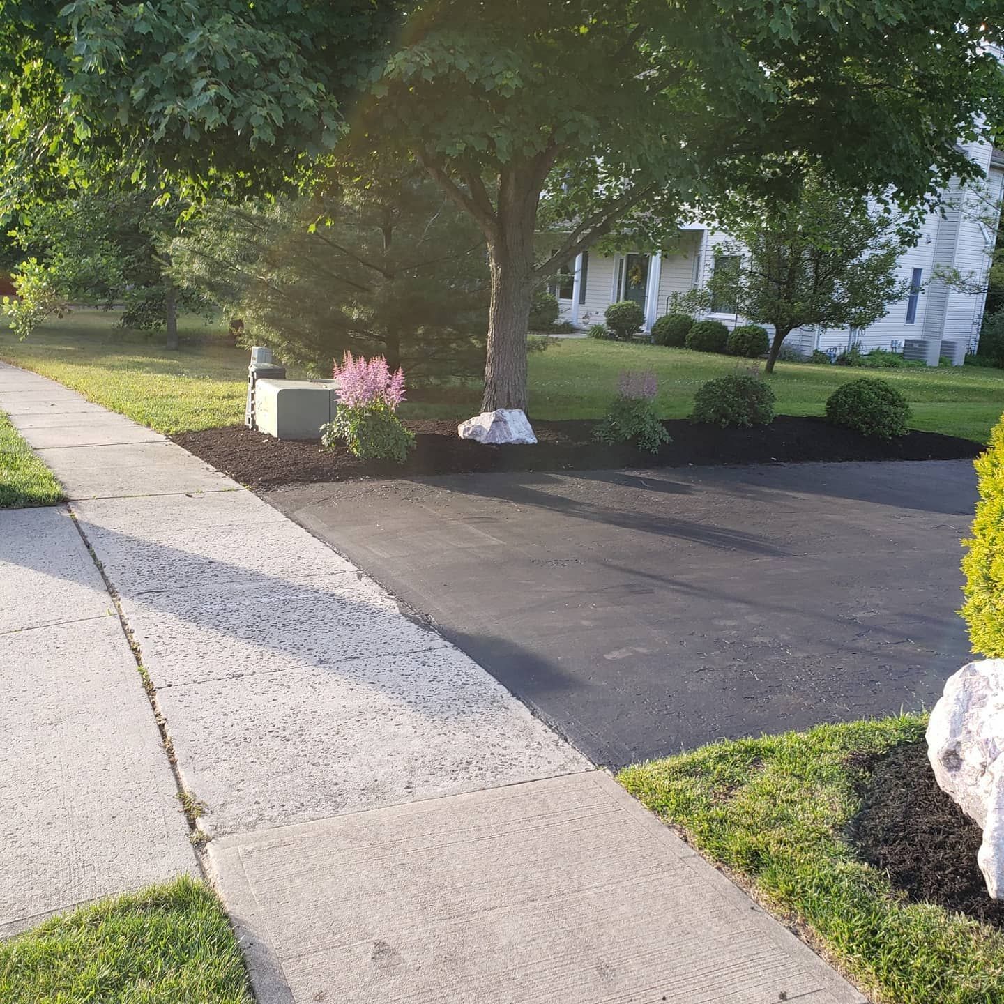 Asphalt driveway next to sidewalk with a landscaped yard and a house in the background.