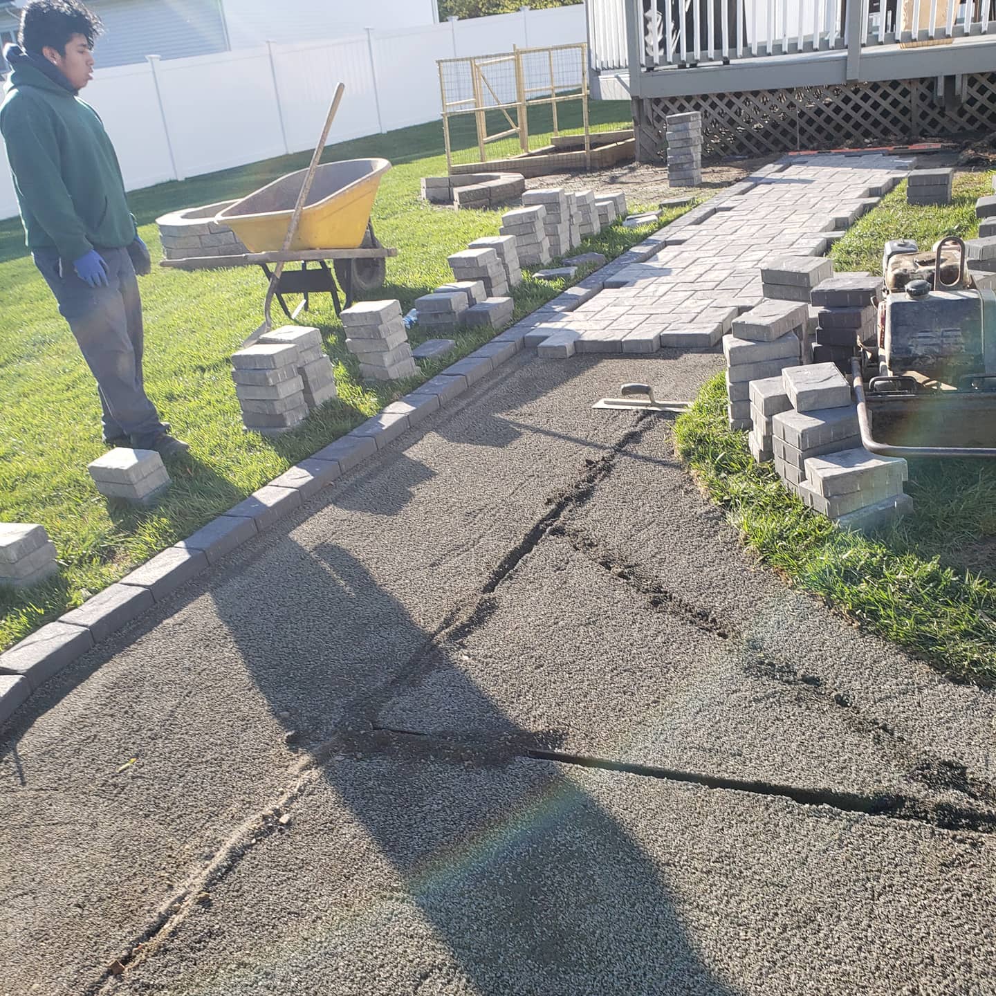 Person laying pavers on a gravel path near a deck, with a wheelbarrow and building materials.