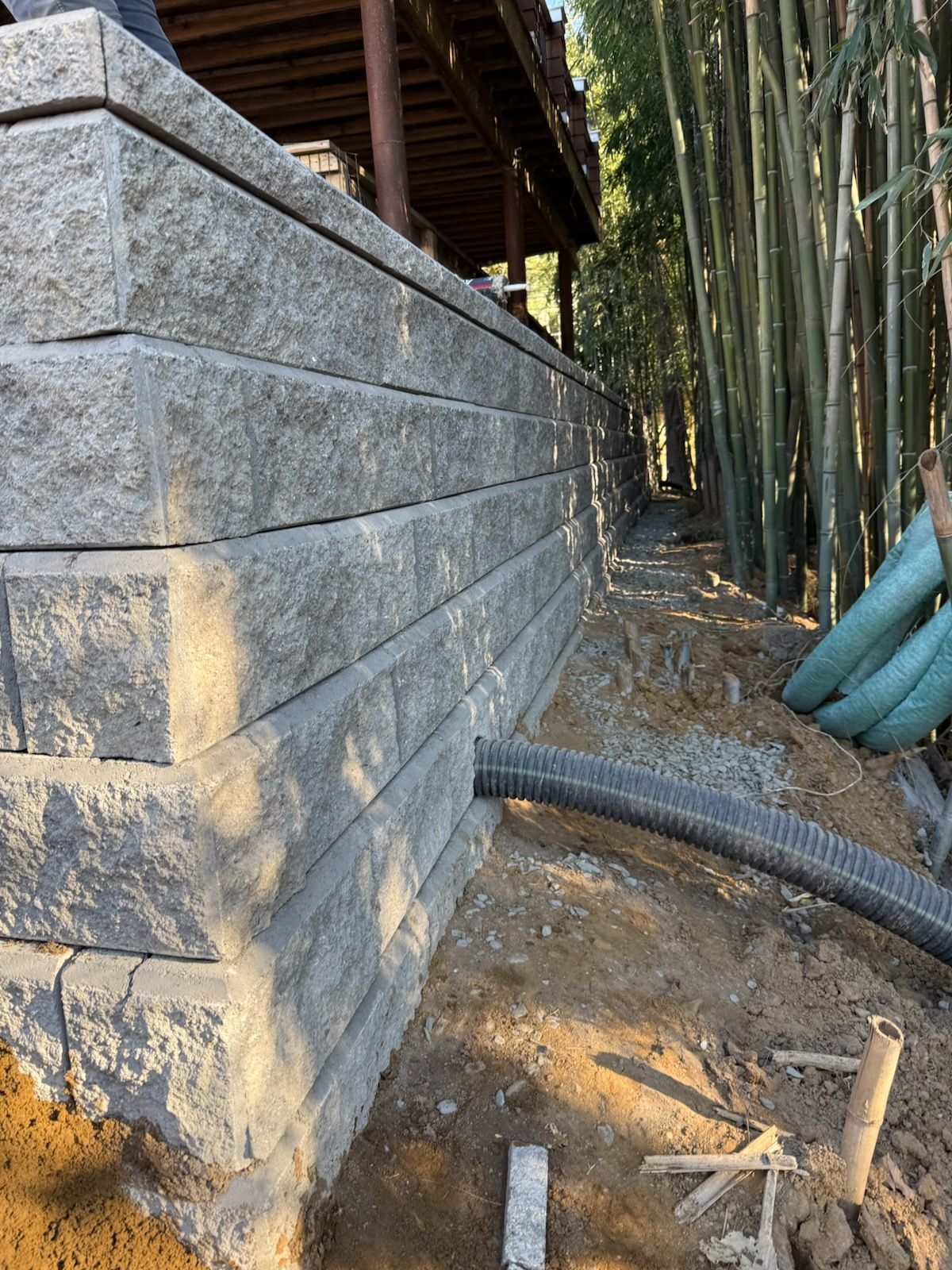 Retaining wall built from gray blocks with a drain pipe, next to a wooded area.