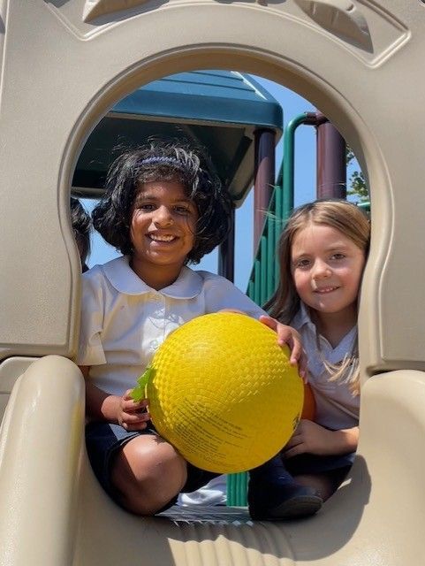 Two girls are sitting on a slide holding a yellow ball