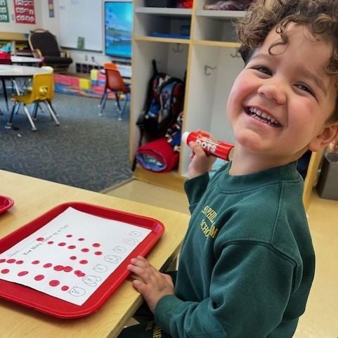 A young boy in a green sweater is holding a red marker and smiling