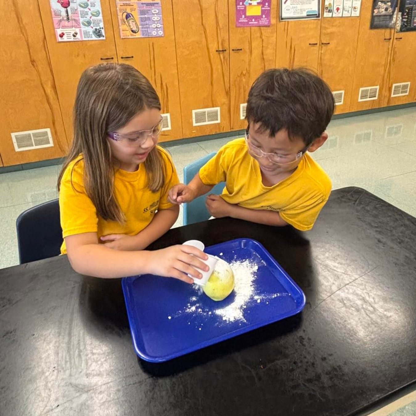 Two children in yellow shirts and safety glasses conduct a science experiment with powder.