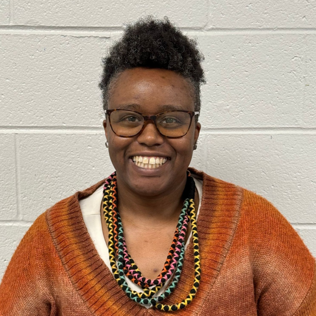 A woman wearing glasses and a sweater is smiling in front of a brick wall.