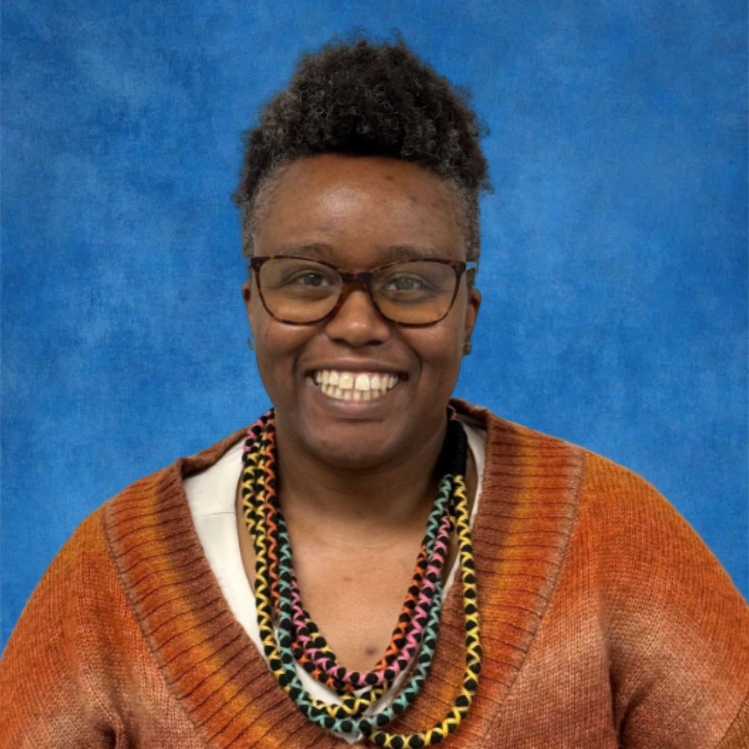 A woman wearing glasses and a sweater is smiling in front of a brick wall.