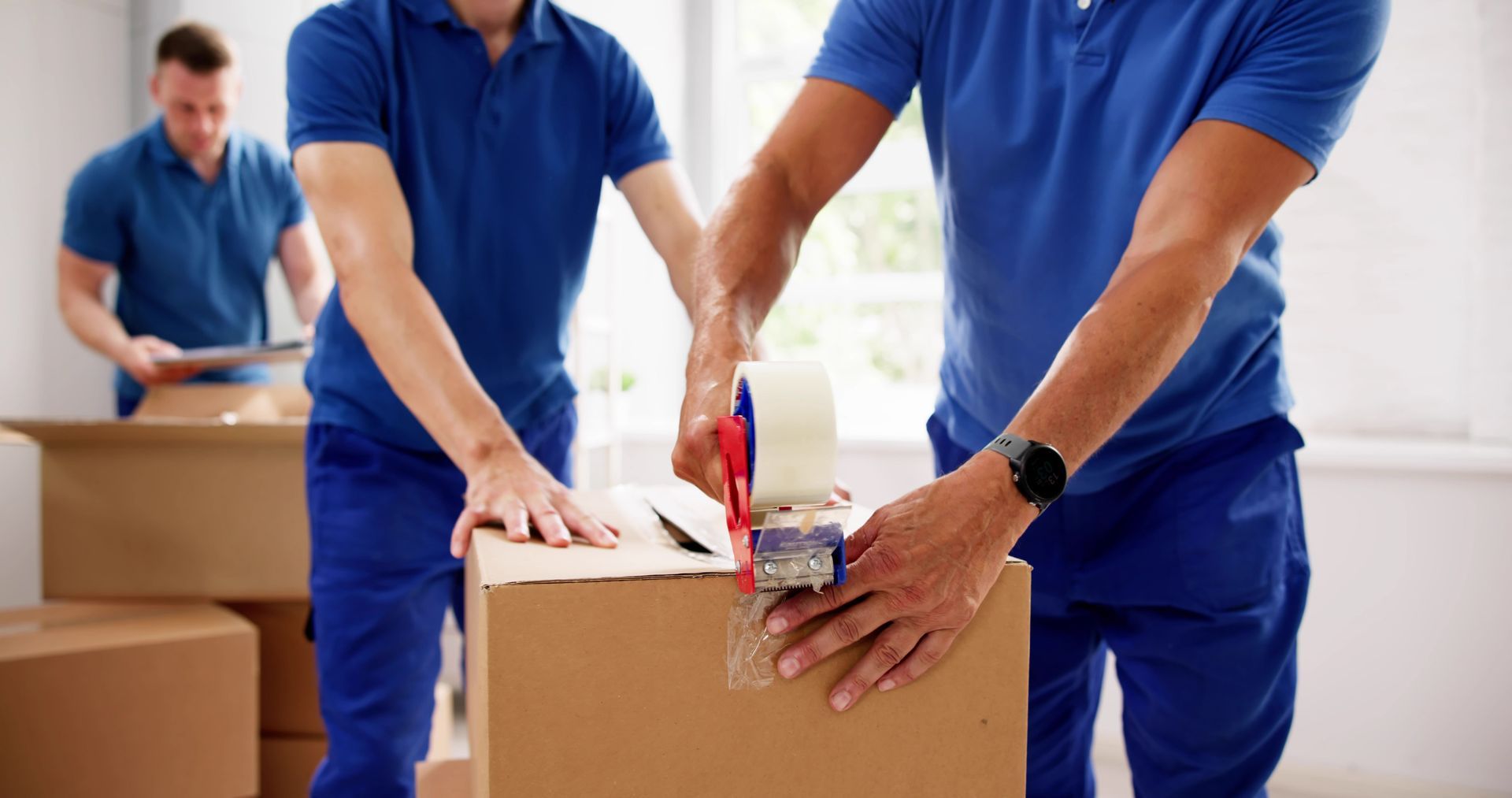 Three people in matching blue uniforms work together to pack and tape cardboard boxes in an indoor setting.