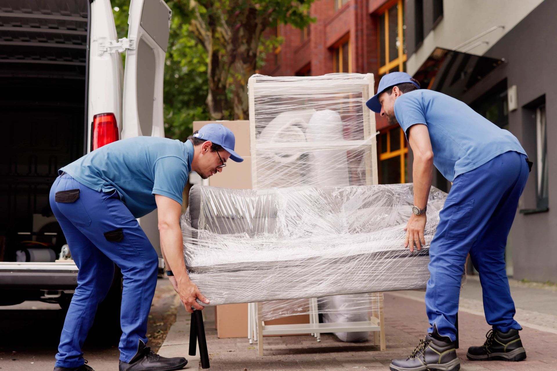 Two movers in blue uniforms carry a sofa wrapped in plastic from a delivery truck parked on a city sidewalk.