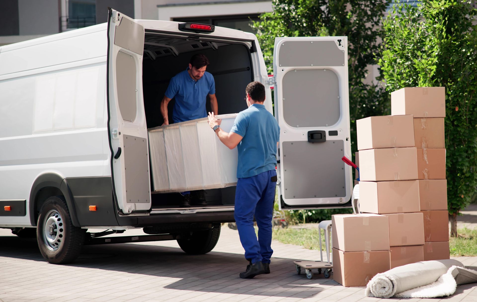 Two people in blue uniforms load a large white appliance into the back of a white delivery van next to stacked boxes.