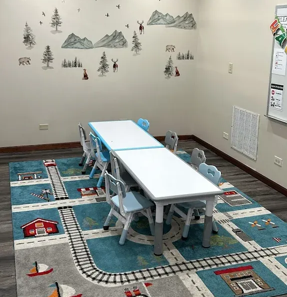 A classroom with a blue rug featuring a road design, tables, chairs, and a mountain scene wall decal.